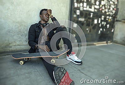 Young depressed and sad black afro American man sitting on grunge street ground corner with skate board feeling depressed Stock Photo