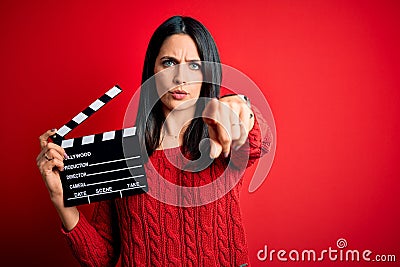 Young director woman with blue eyes making movie holding clapboard over red background pointing with finger to the camera and to Stock Photo