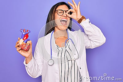 Young doctor cardiology specialist woman holding medical heart over pruple background with happy face smiling doing ok sign with Stock Photo