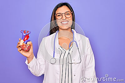 Young doctor cardiology specialist woman holding medical heart over pruple background with a happy face standing and smiling with Stock Photo