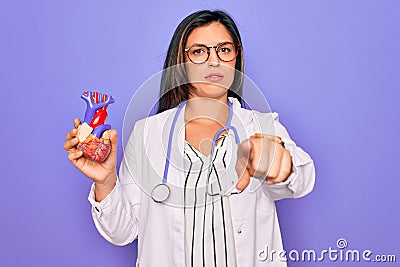 Young doctor cardiology specialist woman holding medical heart over pruple background pointing with finger to the camera and to Stock Photo
