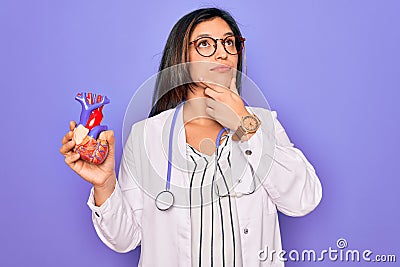 Young doctor cardiology specialist woman holding medical heart over pruple background serious face thinking about question, very Stock Photo