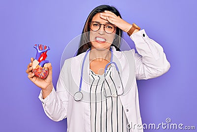 Young doctor cardiology specialist woman holding medical heart over pruple background stressed with hand on head, shocked with Stock Photo