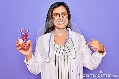 Young doctor cardiology specialist woman holding medical heart over pruple background with surprise face pointing finger to Stock Photo