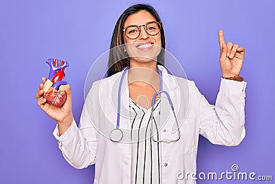 Young doctor cardiology specialist woman holding medical heart over pruple background surprised with an idea or question pointing Stock Photo