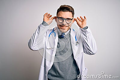 Young doctor man with blue eyes wearing medical coat and stethoscope over isolated background Smiling pulling ears with fingers, Stock Photo