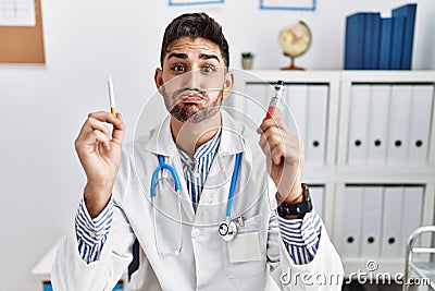 Young doctor man holding electronic cigarette at medical clinic puffing cheeks with funny face Stock Photo