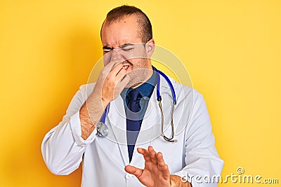 Young doctor man wearing coat and stethoscope standing over isolated yellow background smelling something stinky and disgusting, Stock Photo
