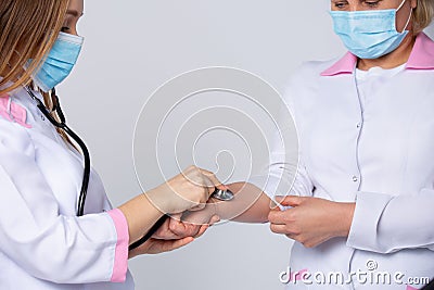 A young doctor in a white lab coat, wearing protective medical mask, is checking the pulse of her colleague- senior Stock Photo