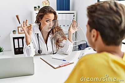 Young doctor woman showing electronic cigarette and normal cigarrete to patient in shock face, looking skeptical and sarcastic, Stock Photo