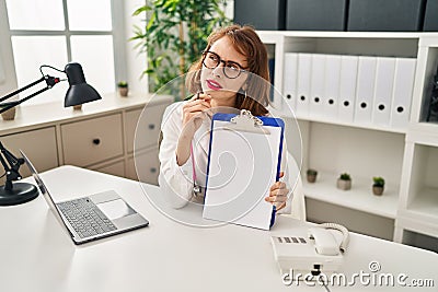 Young doctor woman wearing stethoscope holding clipboard serious face thinking about question with hand on chin, thoughtful about Stock Photo