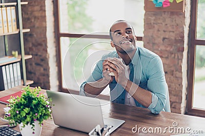 Young dreaming mulatto american worker is thinking in front of laptop at work place. He is happy, smiling, behind him is window an Stock Photo