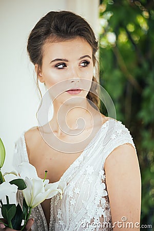 Young elegant woman with wedding bouquet in a light long white dress walking near white house before wedding ceremony. Stock Photo