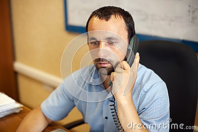 A young employee sits in a chair at a desk in the office and talks on a wired phone. Stock Photo