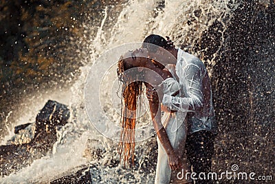 Young enamored couple hugs and kisses under spray of waterfall. Stock Photo