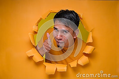 Young entertaining man through torn yellow paper hole showing thump up with facetious of content and happiness Stock Photo