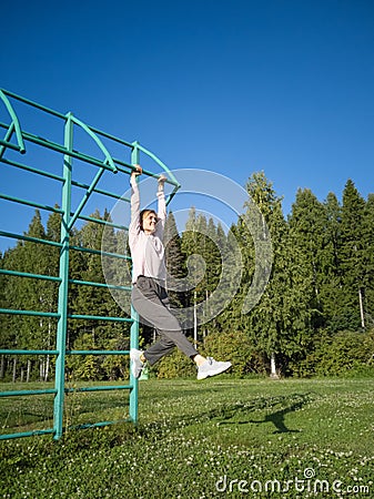 Young European happy girl hanging on the horizontal bar on the street against the background of trees. Stock Photo