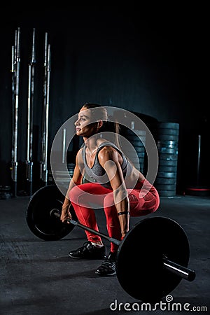 Young, European, muscular girl in red leggings, doing exercise with a barbell in the gym for crossfit. Dark background Stock Photo