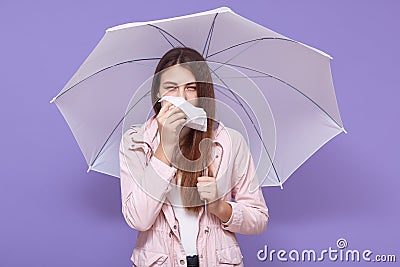 Young European sick young female using paper handkerchief, sneezing, having runny nose, standing under umbrella, feeling unwell, Stock Photo