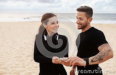 Young european sporty couple working out outdoors at the beach, having break and using mobile phone, standing by seaside Stock Photo