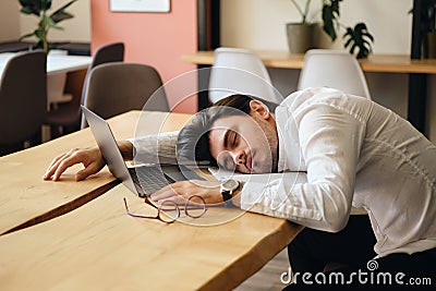 Young exhausted man sitting at the table with laptop while sleeping at work in modern office Stock Photo