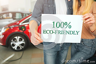 Young family buying first electric car in the showroom. Eco car. Close-up of hands holding paper with word Eco-friendly Stock Photo