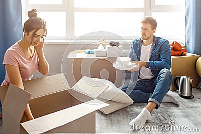 Young family couple bought or rented their first small apartment. Guy sit on floor and hold white plates. Young woman Stock Photo