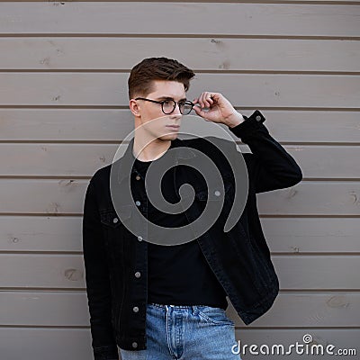 Young fashionable man hipster straightens stylish glasses near wooden wall in street. Handsome guy model in youth black denim Stock Photo