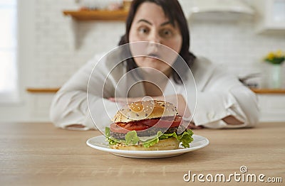 Young fat excited woman sitting at the table in kitchen looking at the plate with big burger. Stock Photo