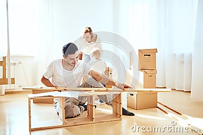 Father assembling furniture in living room of new apartment, mother with son and pile of moving boxes on background Stock Photo