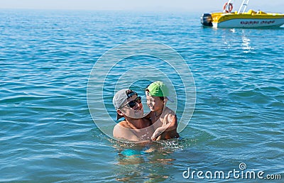 Young father in black sunglasses and smiling baby boy son in green baseball cap playing in the sea in the day time. Positive human Stock Photo