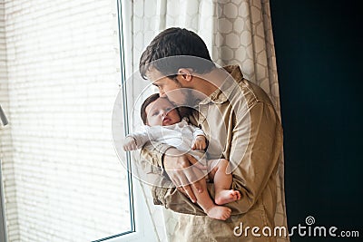 Young father gently holding and kissing his baby near the window at home Stock Photo