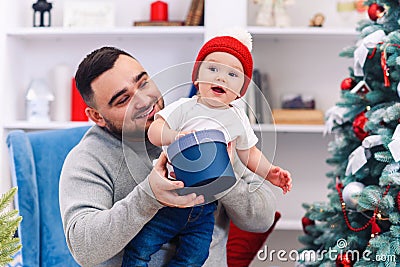 Young father sits in comfortable chair holding amusing toddler and gives gift box to him in the wonderfully decorated Stock Photo