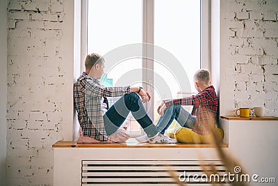 Young father with a son, a boy of 8 years old, sitting on the windowsill and looking out the window, time together, family Stock Photo