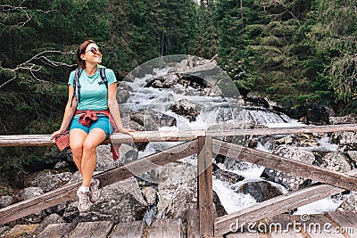 Young female backpacker resting on the mountain cold water stream bridge. Woman sitting on bridge fence smiling and enjoying the Stock Photo