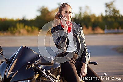 Young female biker with serious expression, has phone conversation via cellular, wears red bandana and leather jacket, poses on mo Stock Photo