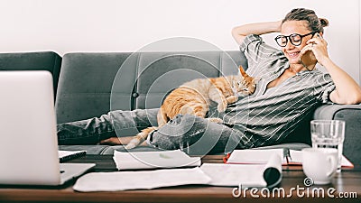 Young female businesswoman student speaks speaks on a mobile phone at home on the couch while working remotely, a ginger cat lies Stock Photo