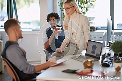 Female colleague sitting on desk, talking to one of the employees, while third one is watching, listening, analyzing information. Stock Photo