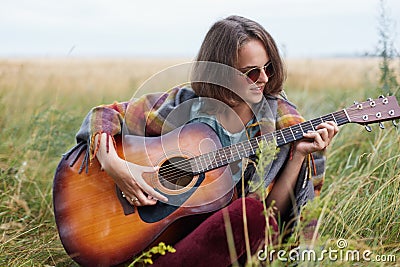 Young female with dark short hair sitting at green grass outdoors playing guitar enjoying nice landscapes singing her favourite so Stock Photo