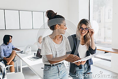 Young female executive explaines new strategy to blonde employee in glasses and smiling. Indoor portrait of Stock Photo