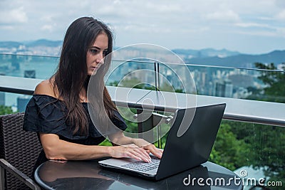 Young female freelancer dressed in black working on a project laptop computer while sitting at rooftop cafe terrace Stock Photo