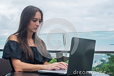 Young female freelancer dressed in black working on a project laptop computer while sitting at rooftop cafe terrace Stock Photo