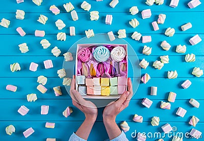 Young female hands holding box full of colorful macaroons and marshmallow with a lot of square different marshmallow on blue Stock Photo