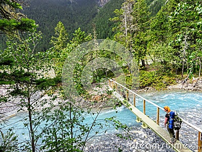 A young female hiker carefully crossing a small skinny wooden bridge over a narrow electric blue rushing river Stock Photo