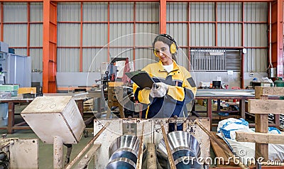 Young female labor dressed in polyester jacket uniform and ear muff reading document on clipboard, stand in front of steel and Stock Photo