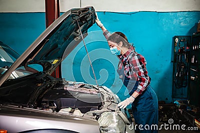 A young female mechanic in a uniform and medical mask opens the hood of a car. Side view. Indoors garage Stock Photo