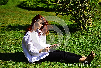Young female poetess with long red hair thinking about read exciting story. Sitting in grass with book. Woman enjoying Stock Photo