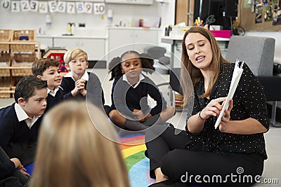 Young female primary school teacher reading a book to children sitting on the floor in a classroom, selective focus Stock Photo