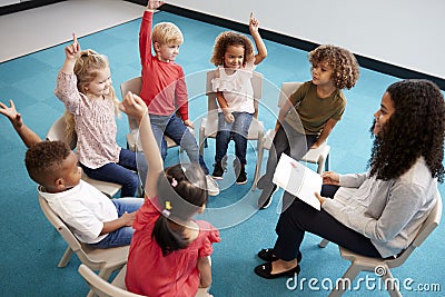 Young female school teacher reading a book to infant school children, sitting on chairs in a circle in the classroom raising hands Stock Photo