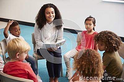 Young female school teacher reading a book to kindergarten children, sitting on chairs in a circle in the classroom listening, clo Stock Photo